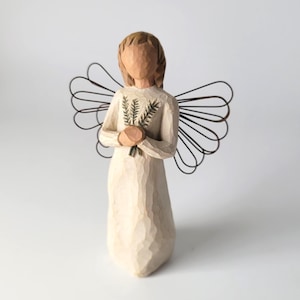 May include: A carved figurine of an angel with a smooth, light cream-colored finish. The figure has a featureless face, short hair, and holds a small bundle of green wheat stalks. Dark brown wire loops form the wings extending from the back.