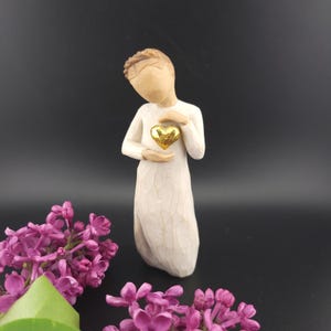 May include: A carved figurine of a person in a long white dress holding a gold heart. The figurine has a neutral skin tone and a simple, serene expression. Purple flowers are in the foreground, with a dark background.