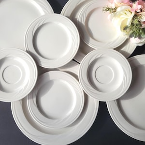 May include: A collection of white ceramic plates and saucers. The plates have a simple, elegant design with a subtle swirl pattern around the rim. The arrangement includes various sizes, suggesting a complete dinnerware set. A few flowers are visible in the background.