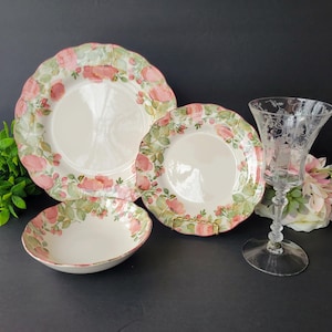 May include: A set of vintage floral dinnerware, including a large plate, a smaller plate, and a bowl, all with a pink rose and green leaf design. A clear etched glass goblet is also present, creating an elegant table setting.