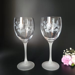 May include: Two clear wine glasses with etched floral and hummingbird designs. The glasses have a frosted stem and base. The background is black.