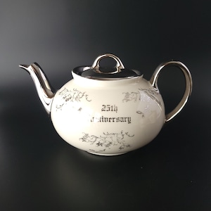 May include: Cream-colored ceramic teapot with silver accents, including the spout, handle, and lid. The teapot is decorated with floral patterns and the text "25th Anniversary".