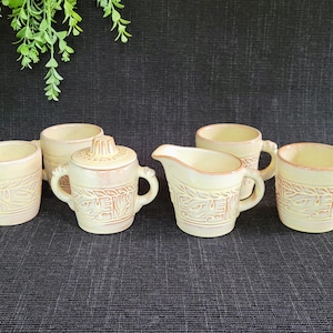 May include: A cream-colored ceramic tea set with embossed leaf and geometric designs. The set includes two mugs, a sugar bowl with a lid, a creamer, and two additional mugs, all with handles.