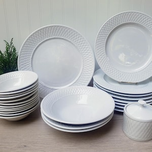 May include: A collection of white ceramic dinnerware, including plates, bowls, and a small lidded container. The plates and bowls have a textured, woven-like border design. The set is arranged on a light-colored wooden surface.
