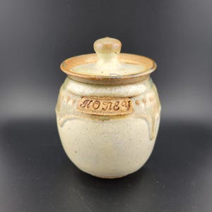 May include: A ceramic honey pot with a lid. The pot is a light beige color with a brown rim and lid. The word "HONEY" is carved into the side of the pot. The pot has a round shape and a small knob on top of the lid.
