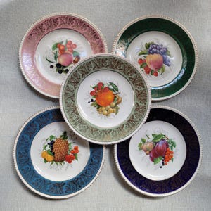 May include: A collection of five decorative plates with fruit illustrations. Each plate features a different fruit arrangement, including peaches, grapes, and plums, set against a white background. The plates have colored rims in pink, green, blue, and gold, with a gold trim.