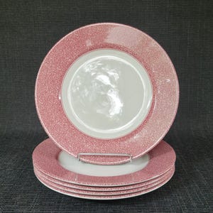 May include: A set of four white and pink patterned dinner plates. The plates have a white center and a pink, textured border. One plate is stacked on top of the others, with a metal plate stand visible.