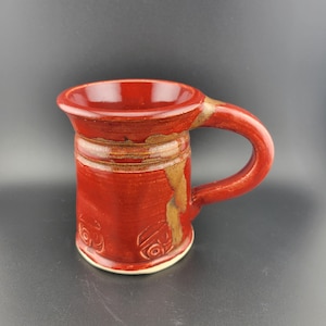 May include: A handmade red ceramic mug with a curved handle. The mug features a wide rim and a cylindrical body with embossed designs. The exterior has a textured, rustic appearance with brown and beige accents. The mug is approximately 4 inches tall.