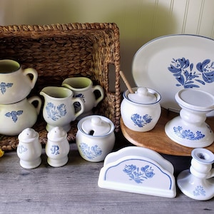 May include: A collection of cream-colored ceramic tableware with blue floral designs. Includes pitchers, salt and pepper shakers, a sugar bowl, a serving platter, a cake stand, a napkin holder, and candle holders. The items are arranged in a wicker basket and on a wooden surface.