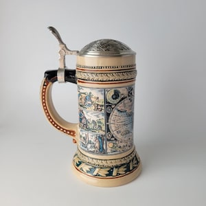 May include: A ceramic beer stein featuring a metallic hinged lid and a curved handle. The body displays a detailed wrap-around illustration with historical scenes and a celestial map. The base and rim are decorated with a dark green leaf pattern.