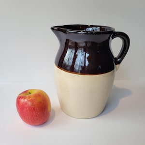 May include: A two-tone ceramic pitcher with a dark brown top and a cream-colored bottom. The pitcher has a handle and a spout. A red and yellow apple sits next to the pitcher.