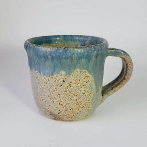 May include: A ceramic mug featuring a speckled, light-colored base with a textured, sandy appearance. The upper portion of the mug is coated in a smooth, glossy blue glaze that drips down over the speckled surface. It has a sturdy handle.