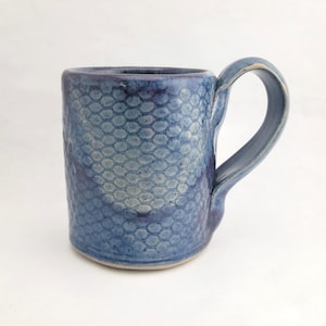 May include: A handmade blue ceramic mug with a textured, honeycomb-like pattern. The mug has a rounded body and a sturdy handle. The color varies from light to dark blue, creating visual interest. A unique piece for enjoying beverages.