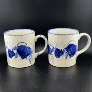 May include: Two white ceramic mugs with blue leaf and vine designs. The mugs have a blue rim and handle. The design is a stylized depiction of leaves and vines, with a whimsical, hand-painted feel. The mugs are set against a dark background.