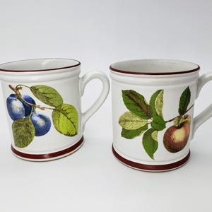 May include: Two white ceramic mugs with brown trim. One mug features a blue plum branch with green leaves. The other mug features a red apple branch with green leaves.