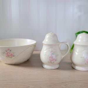 May include: A cream-colored ceramic bowl and two salt and pepper shakers. Each piece is decorated with a pink rose design. The shakers have handles and a perforated top. The bowl has a diamond pattern.