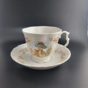 May include: A white porcelain teacup and saucer set featuring a scalloped edge and a decorative handle. The cup displays a colorful illustration of a rabbit wearing a green hat, surrounded by delicate floral and botanical motifs in soft tones.