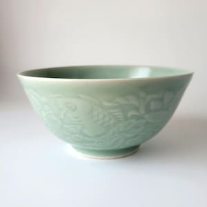 May include: A pale celadon green ceramic bowl featuring a relief design of a fish swimming among aquatic leaves. The bowl has a smooth, glossy finish and a rounded shape with a slightly flared rim, resting on a small, circular footed base.