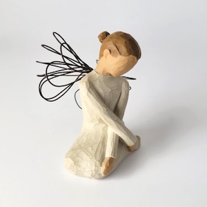 May include: A carved figurine of a seated figure with a smooth, light cream-colored dress and brown hair pulled back into a bun. The figure features dark, thin, wire-like wings extending from the back, with a simple, faceless, and minimalist aesthetic.