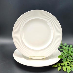 May include: Two round white ceramic plates featuring a smooth center and a wide, textured rim with vertical ribbed detailing. The plates are stacked, with the top plate held upright by a small gold-toned metal stand, showcasing the design.