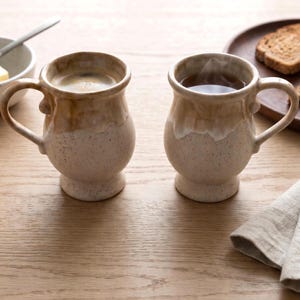 May include: Two ceramic mugs with a rounded base and a pedestal foot. The mugs feature a speckled cream-colored lower body and a smooth, glossy tan glaze on the upper portion and handle. Each mug is filled with a dark liquid, one showing visible steam.