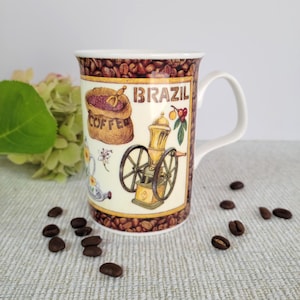 May include: White ceramic coffee mug with a Brazil theme. The mug features illustrations of coffee beans, a coffee grinder, and a bag of coffee beans. The word "BRAZIL" is printed in brown. Coffee beans are scattered around the mug.