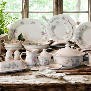 May include: A collection of white porcelain dinnerware with a floral design, including plates, bowls, cups, and serving dishes. The floral pattern features purple and green accents. The set is arranged on a rustic wooden table.