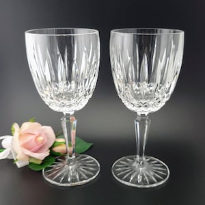 May include: Two clear crystal wine glasses with intricate vertical and diamond-shaped cuts. The glasses have a classic design with a round base and a long stem. A pink rose with green leaves is in the background. The glasses are on a black surface.
