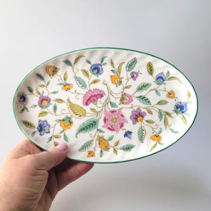 May include: An oval ceramic serving dish with a scalloped edge and a thin green rim. The surface features a colorful botanical pattern with pink, blue, yellow, and purple flowers, green leaves, and brown stems on a white background.