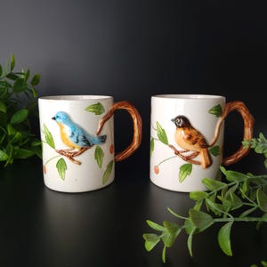 May include: Two white ceramic mugs with 3D bird designs. One mug features a blue and yellow bird, the other a brown and orange bird. Both have branch-shaped handles and are decorated with green leaves and red berries.