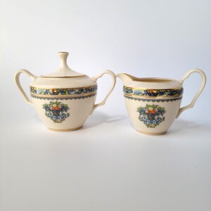 May include: Cream-colored sugar bowl with lid and creamer set. Both pieces feature a gold rim and a blue and gold decorative band. A floral basket design is centered on each piece, adding a touch of elegance to this vintage tableware.