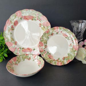 May include: A set of floral-patterned dinnerware featuring a large plate, a smaller plate, and a bowl. The plates and bowl have a white background with pink roses and green leaves around the edges. A clear glass vase is in the background.