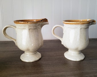 May include: Two cream-colored ceramic creamer pitchers with brown rims and a textured surface. The pitchers have a footed base and a handle.