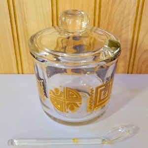 May include: A clear glass jar with a lid and a small spoon. The jar has a gold geometric pattern around the sides. The spoon is clear and rests beside the jar. The jar and spoon are on a white surface.