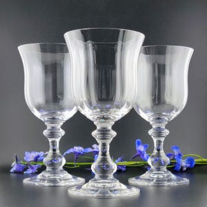 May include: Three clear, stemmed wine glasses with a tulip-shaped bowl and ornate stem. The glasses are arranged in a group, with blue flowers at the base. The glasses are made of clear glass.