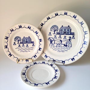 May include: A set of three white ceramic plates with blue decorative borders. The plates feature blue illustrations of a house, tree, and farming scene. The plates are displayed on gold and silver plate stands.