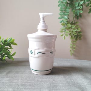 May include: A white ceramic soap dispenser with a pump top. The dispenser has a cylindrical body with a decorative band featuring green floral accents and a green stripe at the base. The pump is white.