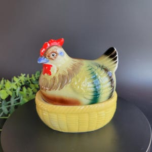 May include: A ceramic hen-shaped container with a yellow basket base. The hen is painted with brown, green, and yellow hues, featuring a red comb and wattle, and a blue beak. The container is designed to hold eggs, adding a touch of farmhouse charm.