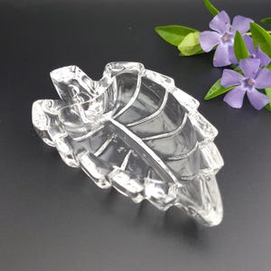 May include: A clear glass dish shaped like a leaf with defined veins and a scalloped edge. The dish has a smooth, reflective surface and a pointed tip. It is positioned near a small cluster of purple periwinkle flowers with green leaves.