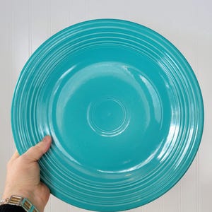 May include: A large, turquoise-colored ceramic plate with concentric ridged detailing. The plate is round and has a glossy finish. The plate is being held by a person. The background is a white vertical panel.