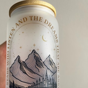 Op de afbeelding: Een witte, gematteerde glazen pot met een gouden deksel en een gouden ontwerp. Het ontwerp toont een skyline met bergen op de achtergrond, sterren en een halve maan. De tekst luidt "To the stars who listen and the dreams that are answered Velaris + City of Starlight +"