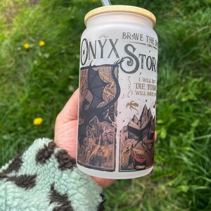 Peut inclure: Un gobelet en verre d&eacute;poli avec un couvercle en bambou et une paille transparente. Le gobelet pr&eacute;sente les mots "Onyx Storm" et un graphisme d'un dragon et d'un livre. Le texte "Brave the Dark" et "I will not die today, I will save them" sont &eacute;galement visibles.