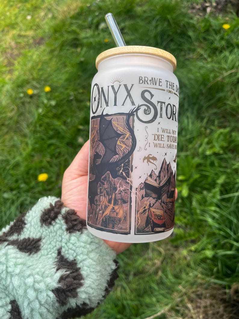 Peut inclure: Un gobelet en verre d&eacute;poli avec un couvercle en bambou et une paille transparente. Le gobelet pr&eacute;sente les mots "Onyx Storm" et un graphisme d'un dragon et d'un livre. Le texte "Brave the Dark" et "I will not die today, I will save them" sont &eacute;galement visibles.