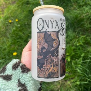 Peut inclure: Un gobelet en verre d&eacute;poli avec un couvercle en bambou et une paille transparente. Le gobelet pr&eacute;sente le texte "Onyx Storm" et un dessin d'un dragon noir au-dessus d'un ch&acirc;teau. L'arri&egrave;re-plan est de l'herbe verte.