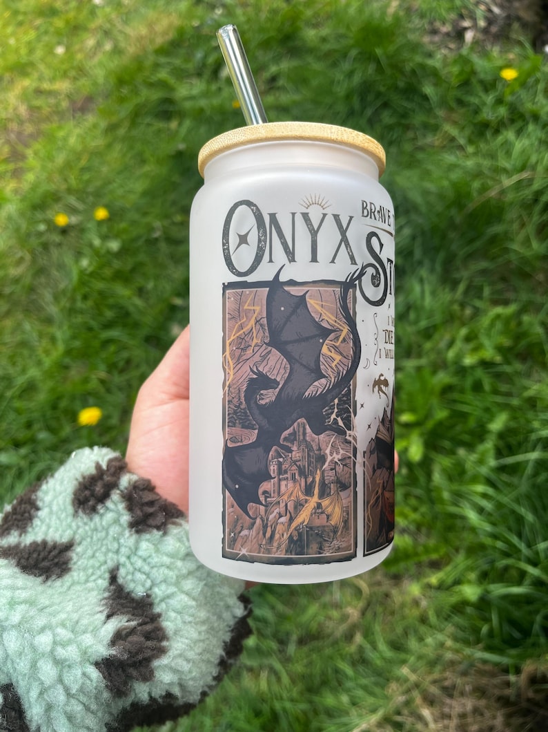 Peut inclure: Un gobelet en verre d&eacute;poli avec un couvercle en bambou et une paille transparente. Le gobelet pr&eacute;sente le texte "Onyx Storm" et un dessin d'un dragon noir au-dessus d'un ch&acirc;teau. L'arri&egrave;re-plan est de l'herbe verte.