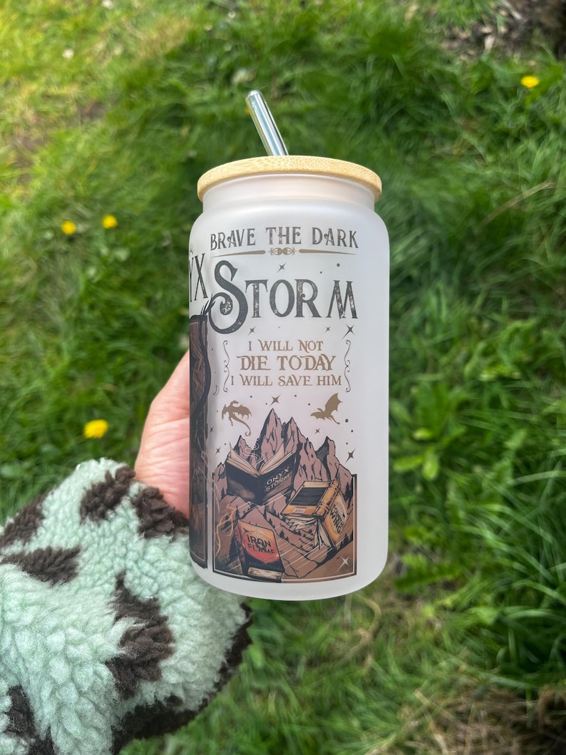 Peut inclure: Un gobelet en verre d&eacute;poli avec un couvercle en bambou et une paille argent&eacute;e. Le gobelet pr&eacute;sente le texte "Brave the Dark" et "Storm" avec un graphique de montagne et de livre. Le design comprend le texte "I will not die today, I will save him."