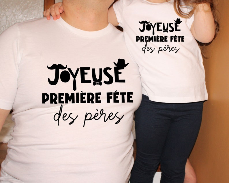 K&ouml;nnte beinhalten: Zwei wei&szlig;e T-Shirts mit schwarzem Text, der "Joyeuse Premiere Fete des Peres" lautet. Der Text ist auf Franz&ouml;sisch und bedeutet "Frohes erstes Vaterfest".