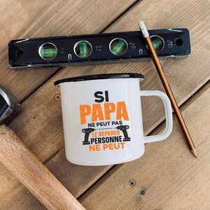 K&ouml;nnte beinhalten: Wei&szlig;e Email Tasse mit schwarzem Rand und Henkel. Die Tasse hat einen orangen Text, der "Si Papa ne peut pas le reparer personne ne peut" lautet. Auf der Tasse sind zwei schwarze und orangefarbene Bohrer abgebildet.