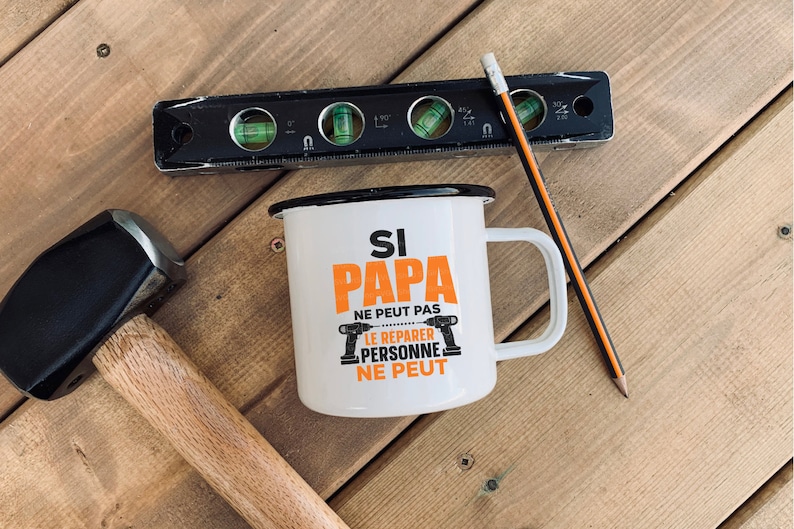 K&ouml;nnte beinhalten: Wei&szlig;e Email Tasse mit schwarzem Rand und Henkel. Die Tasse hat einen orangen Text, der "Si Papa ne peut pas le reparer personne ne peut" lautet. Auf der Tasse sind zwei schwarze und orangefarbene Bohrer abgebildet.