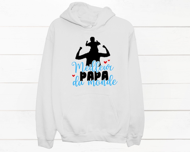 K&ouml;nnte beinhalten: Wei&szlig;er Hoodie mit einer schwarzen Silhouette eines Vaters, der ein Kind auf seinen Schultern tr&auml;gt. Der Text "Meilleur Papa du monde" ist in Blau und Rot auf dem Hoodie gedruckt.